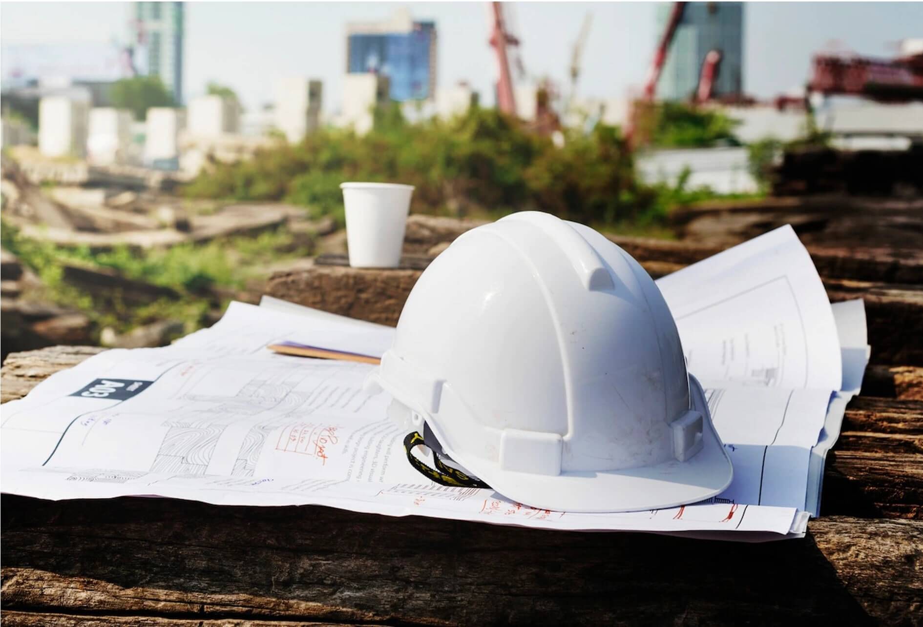 Maintenance et travaux tous corps d’état Au 1er plan, un casque sur des plans d’architecte avec un gobelet, sur fond de friche de chantier