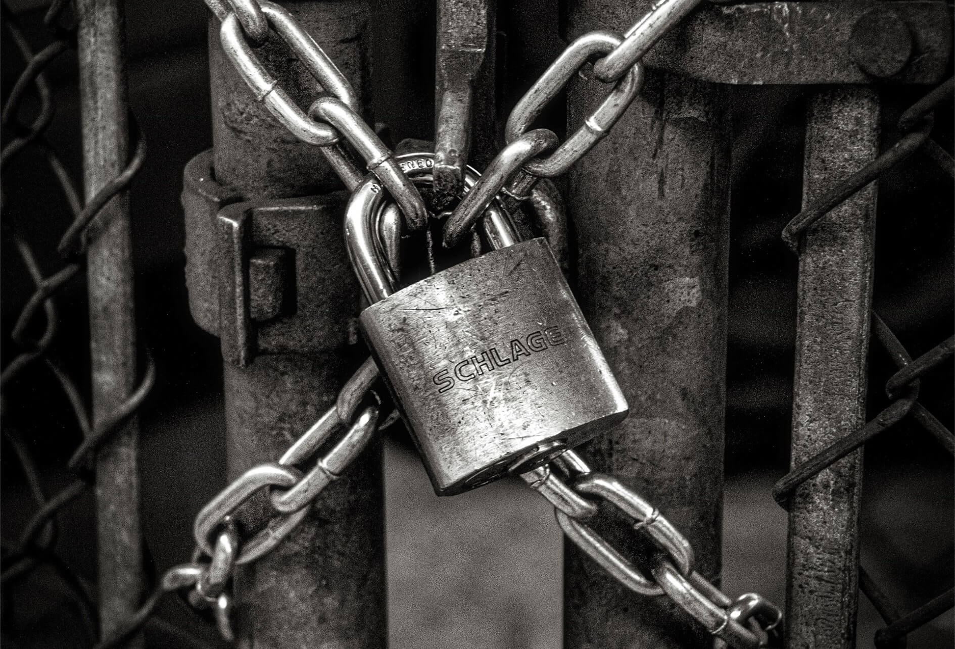 Assistance technique appel d’offres sécurité En noir et blanc, un portail grillagé verrouillé par un cadenas et des chaînes.