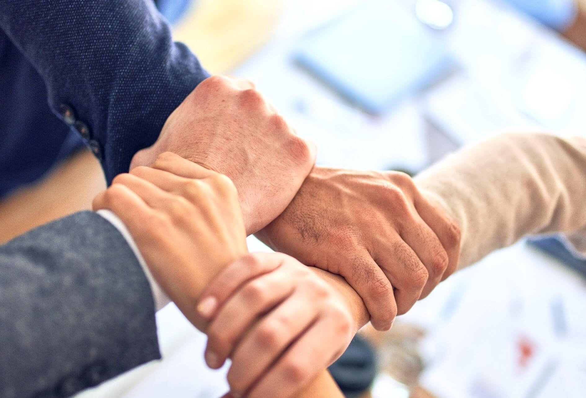 Entrecroisement de 4 mains se tenant solidairement les poignets sur fond flou de bureau