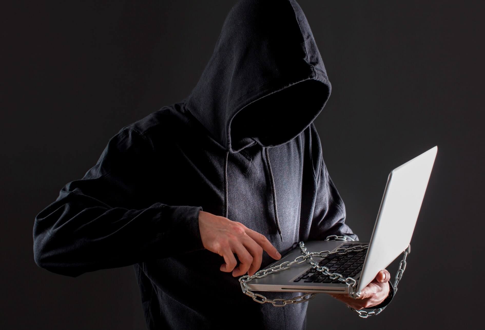 Cyberdéfense et protection des données Sur fond sombre, un pirate avec capuche manipule un ordinateur couvert de chaînes.